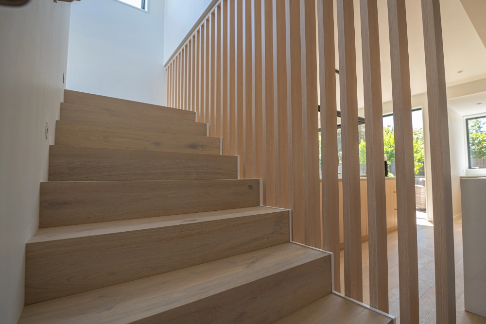 Modern indoor wooden staircase with vertical slat railing beside bright living area windows.
