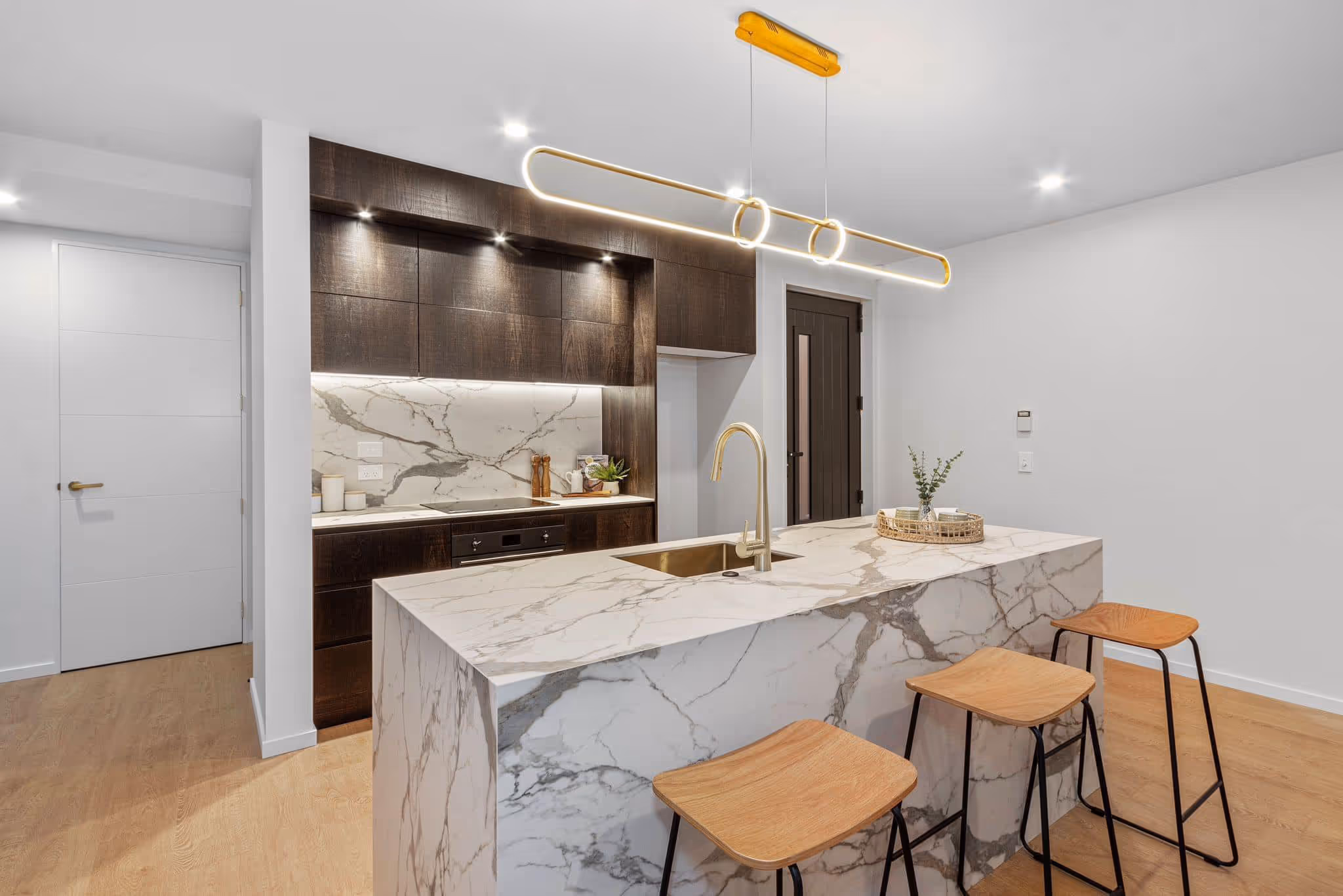 Modern kitchen with marble island, three wooden stools, dark cabinets, and gold-finished faucet and light fixture.