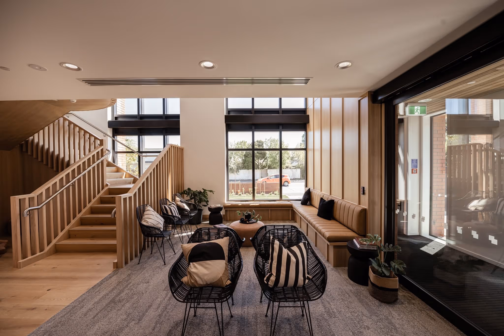 Modern lounge with black chairs, wooden staircase, long bench, and large windows showing outside view.