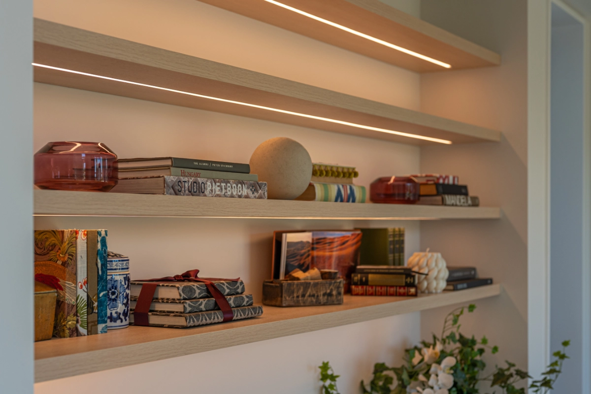 Two wooden shelves with books, decorative objects, and soft LED lighting on a beige wall.