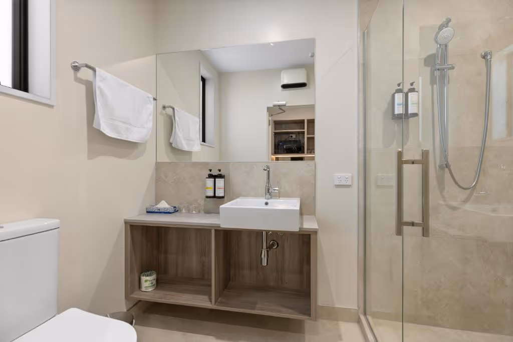 Modern bathroom with wall-mounted sink, glass shower, white towels, and beige walls and floor.