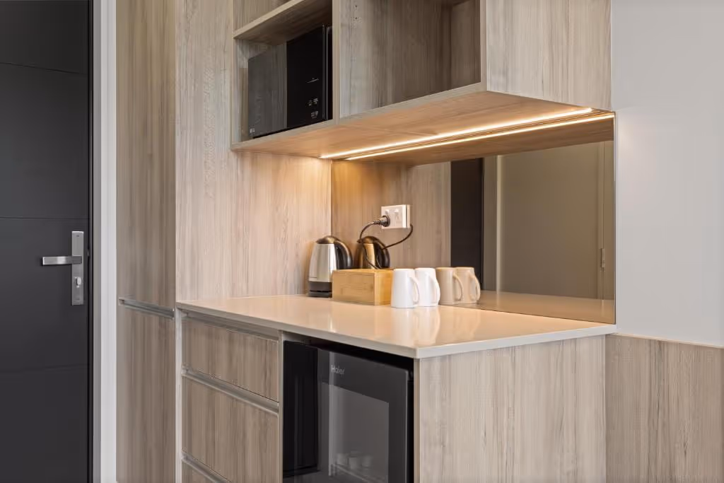 Modern kitchenette with wooden cabinets, a kettle, teapot, mugs, and a mini fridge under the countertop.