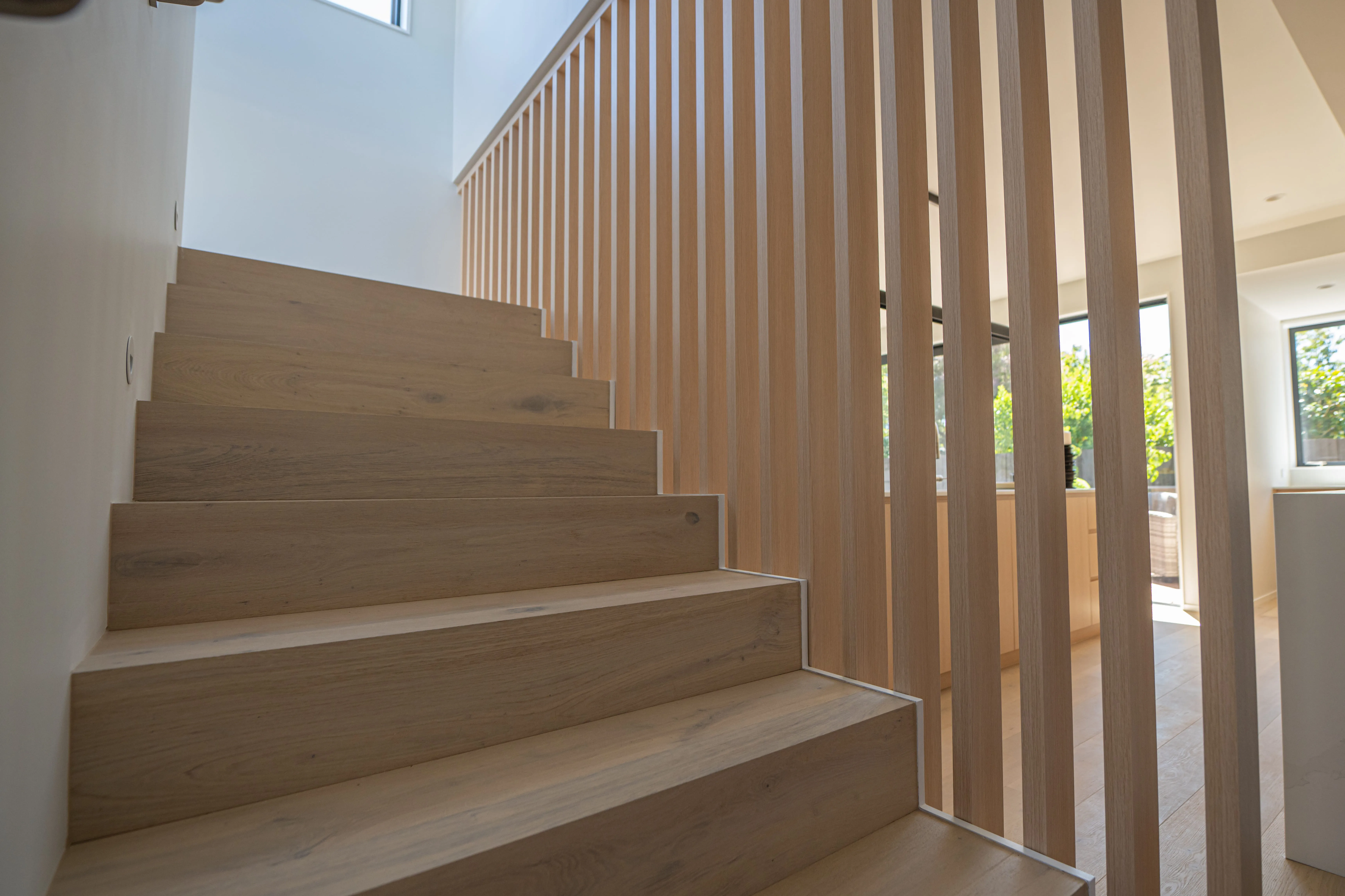 Wooden staircase with vertical slat railing beside bright modern living space with large windows.