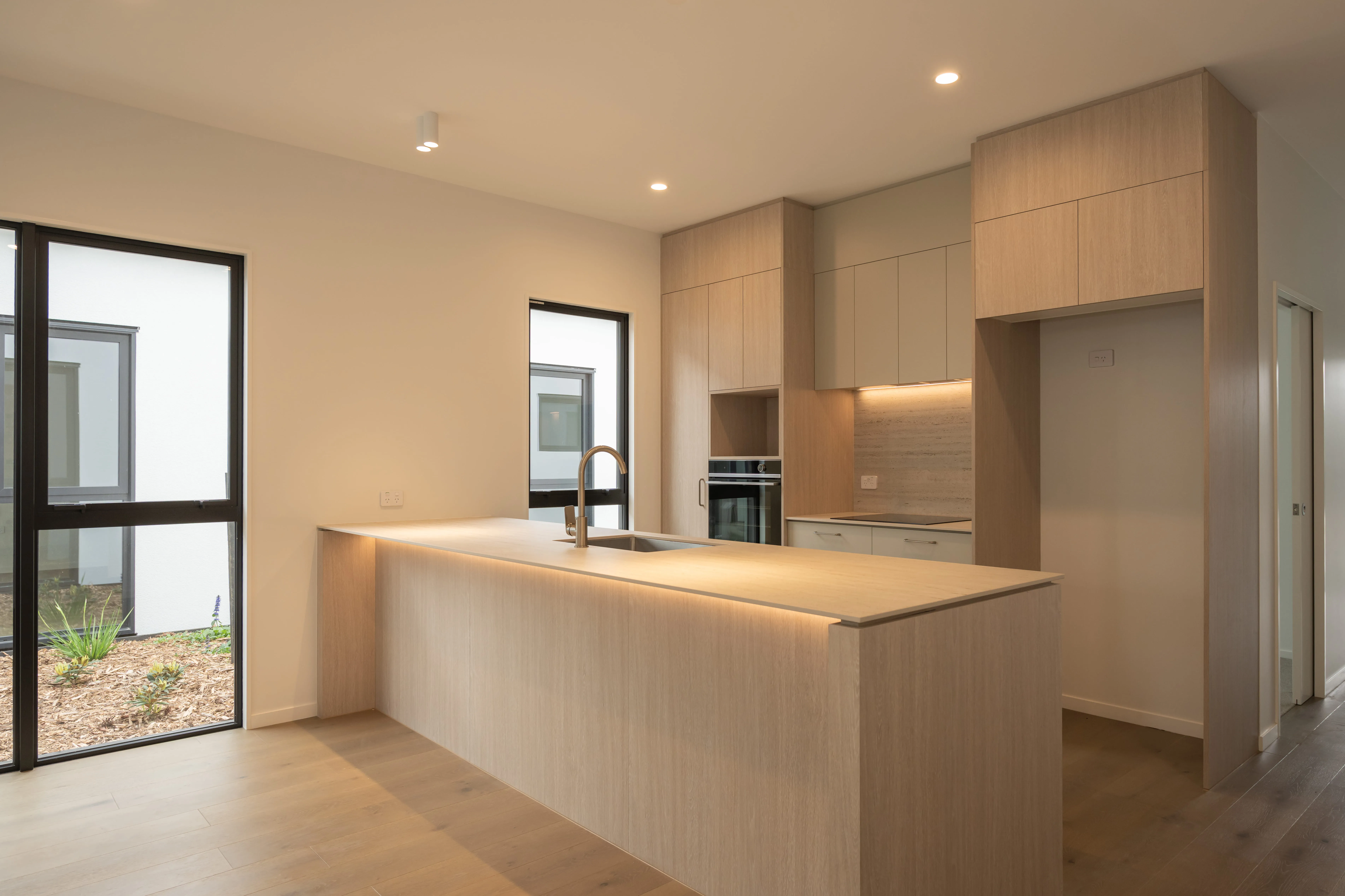 Modern kitchen with wood cabinetry, long island with sink, built-in oven, and large windows.