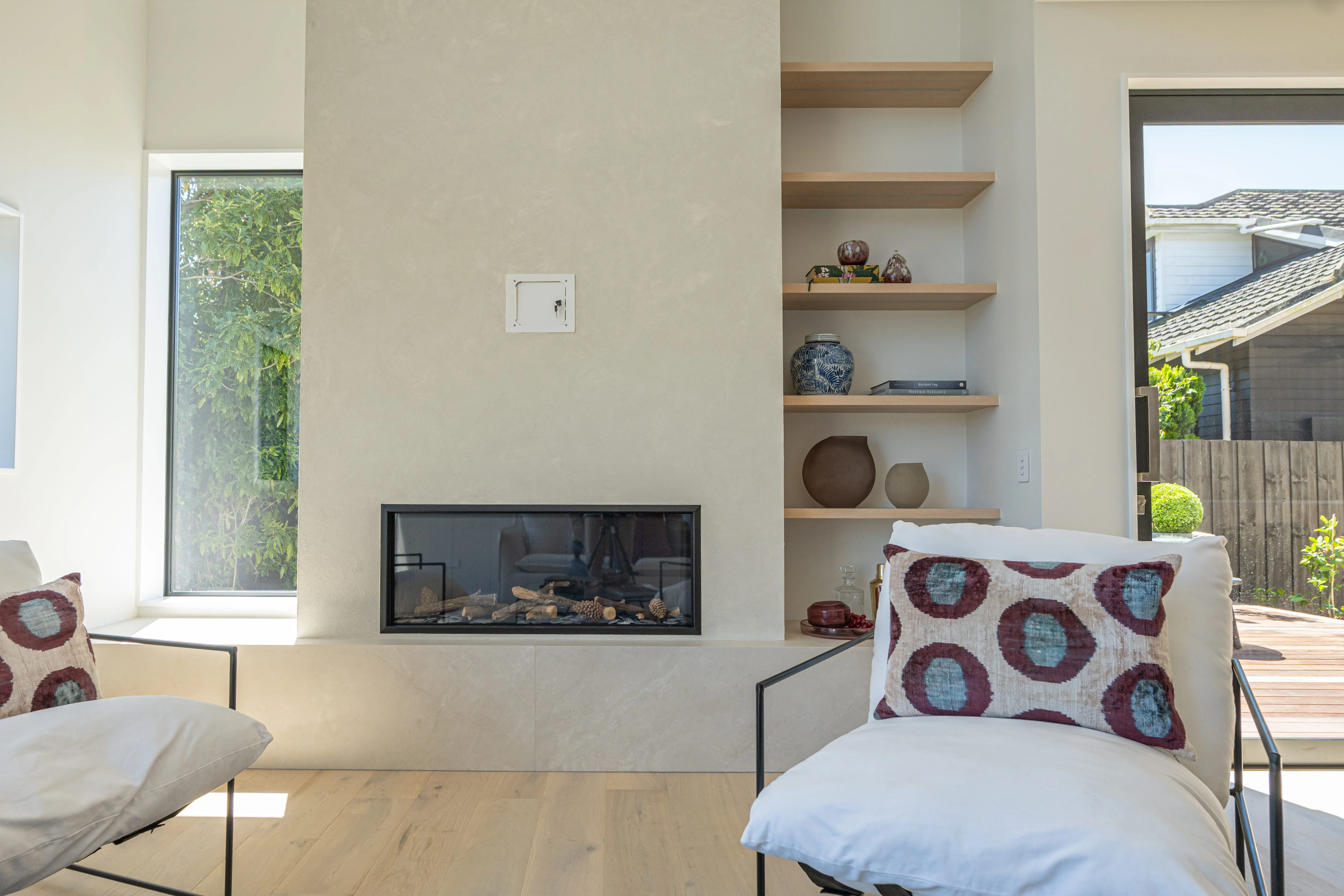 Modern living room with built-in gas fireplace, two white cushioned chairs, and wooden shelves with decor.