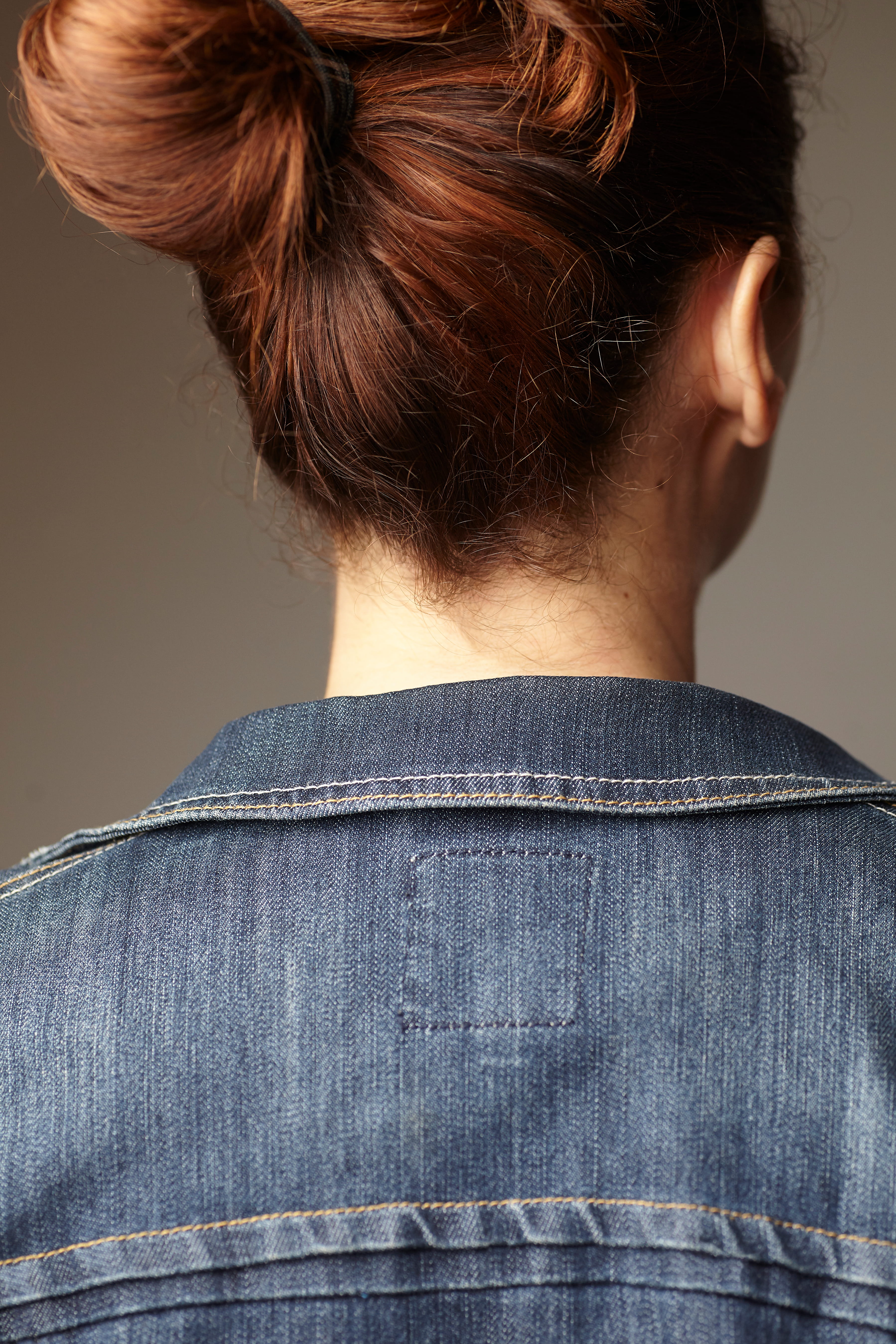 Back view of person with red hair in a bun wearing a blue denim jacket with visible stitching.