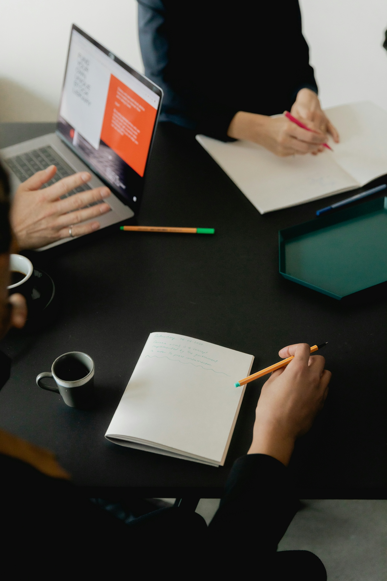 People working at a table with notebooks, pencils, a laptop, and coffee cups.