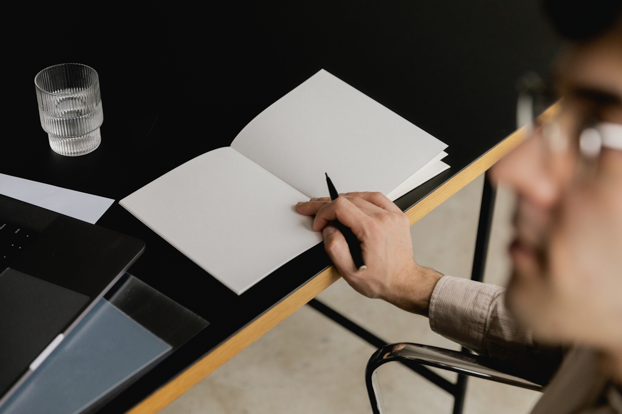 Person holding pen over a blank open notebook on a black table with a glass of water nearby.