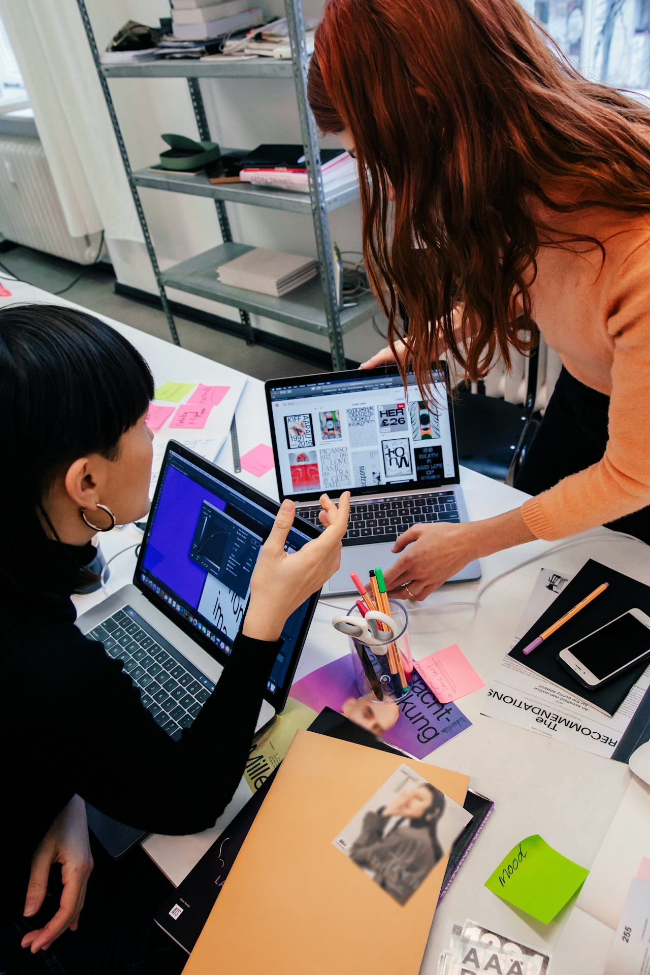 Two women collaborate with laptops open on a desk covered in papers, pens, and sticky notes.