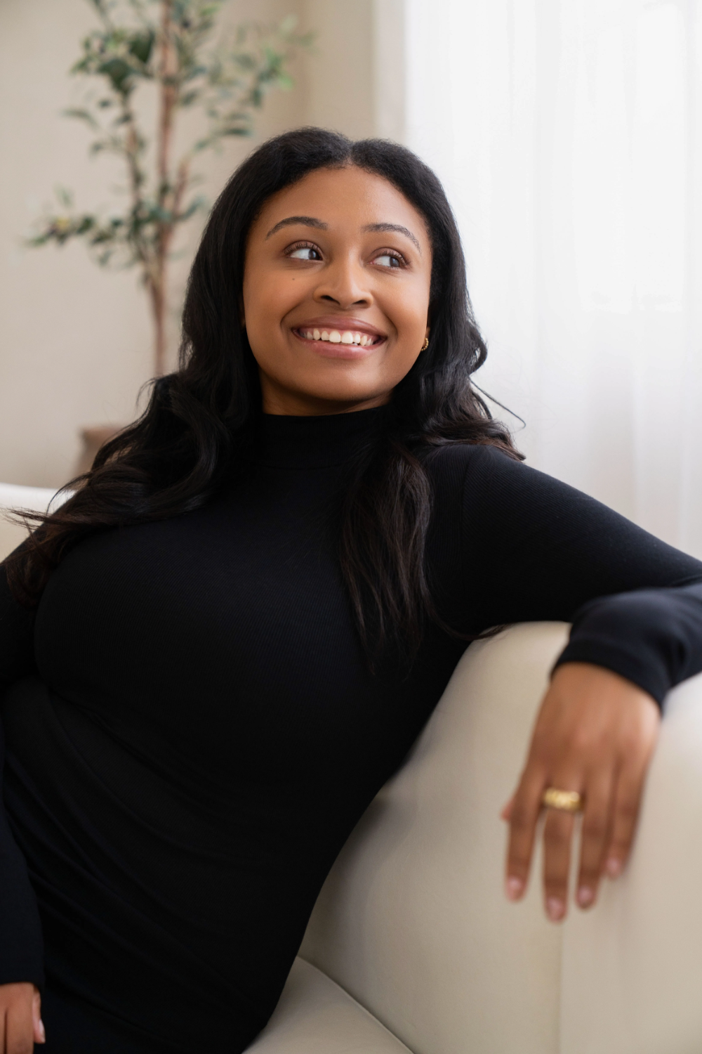 Smiling woman with long dark hair in black top sitting on a light sofa indoors.