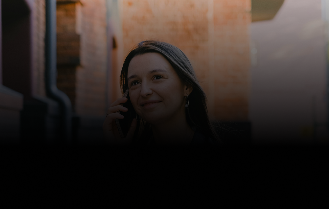 Young woman with long hair smiling and talking on a smartphone outdoors near brick walls.
