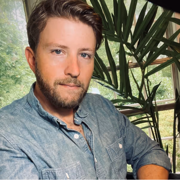 Bearded man wearing a denim shirt sitting indoors near a large green plant and windows.
