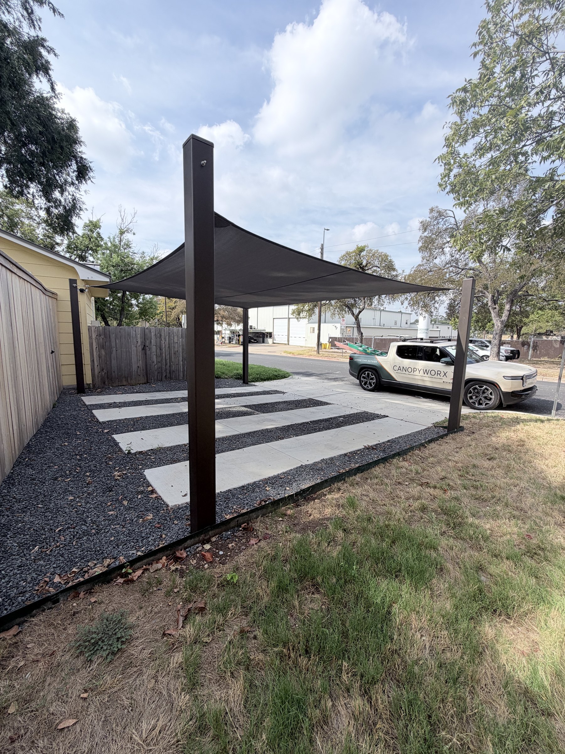 Four-corner carport sail on dark steel posts over a striped concrete-and-gravel pad