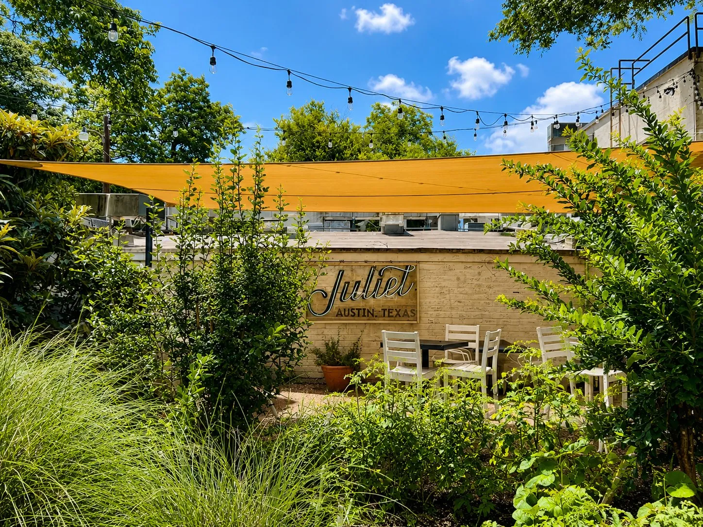 Juliet's garden patio seen through the plantings, with the yellow sail and string lights overhead