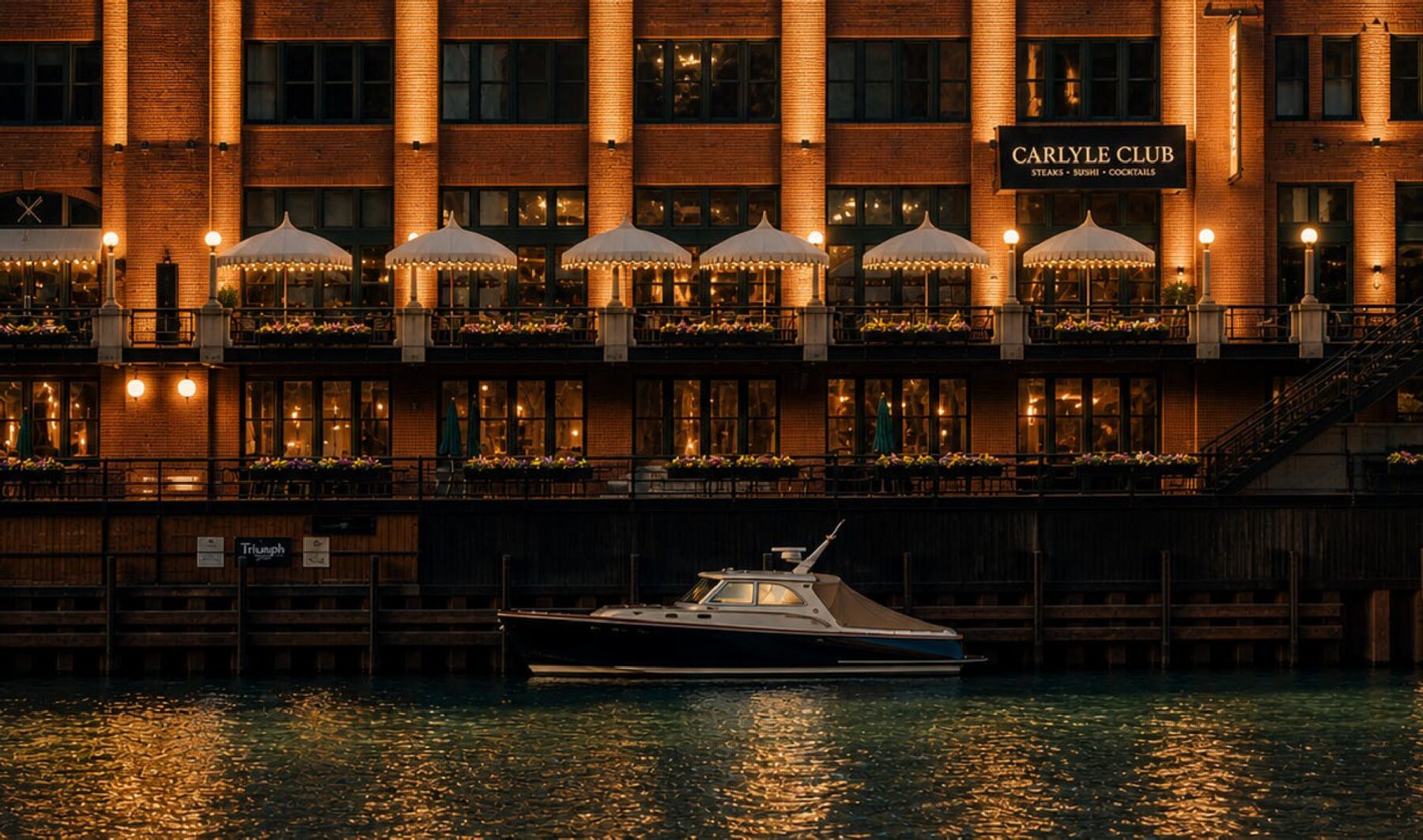 The water-front patio seen from the river at dusk, umbrellas lit beneath the Carlyle Club sign
