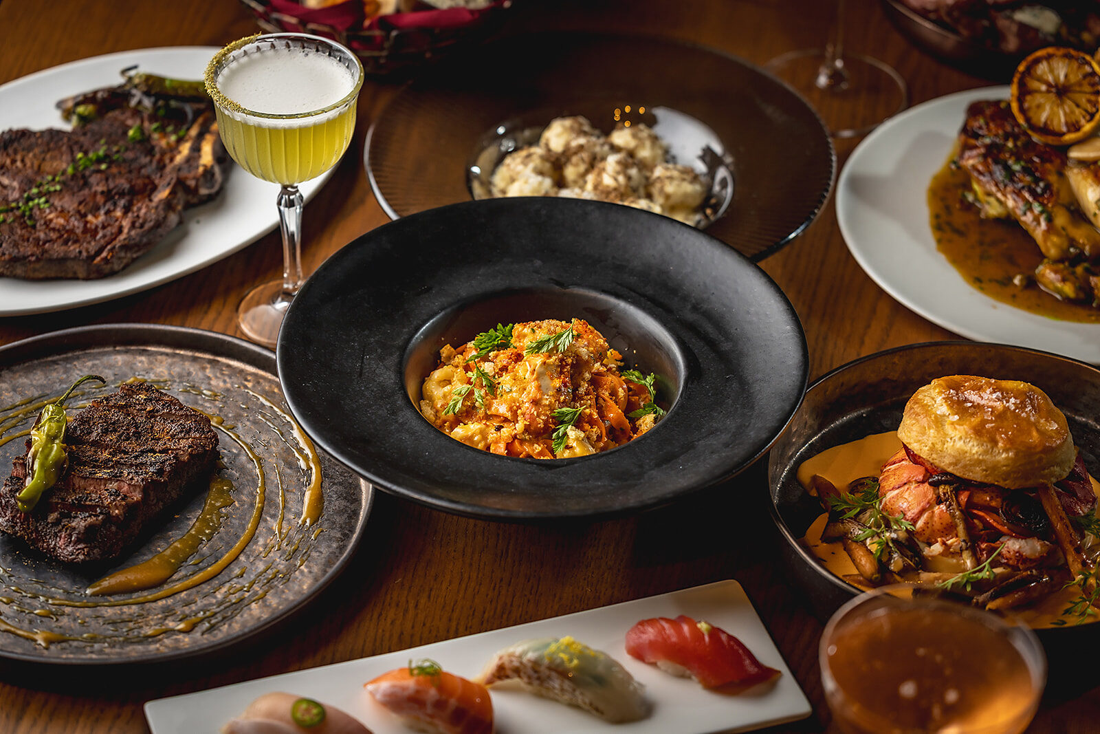 A table set with a dry-aged ribeye, nigiri, handmade pasta, lobster pie, and a cocktail
