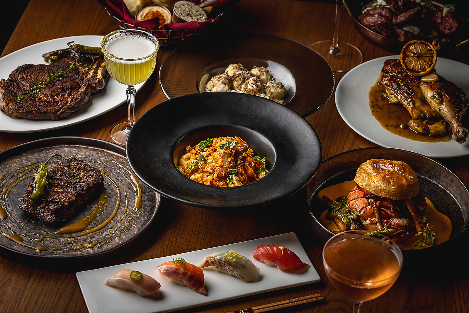 A table set with steaks, nigiri, handmade pasta, roast chicken, lobster pie, and cocktails