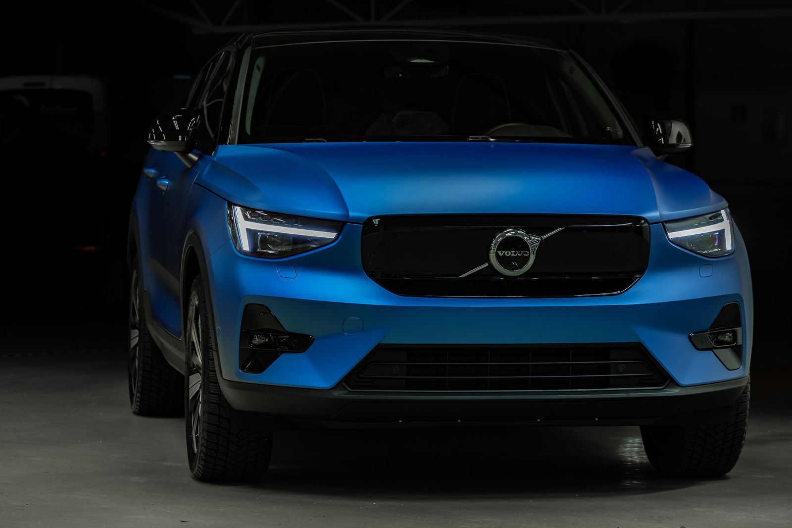 Front view of a blue Volvo car with illuminated headlights in a dark setting.