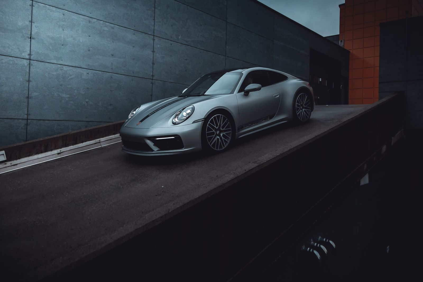 Sølv Porsche sportsbil parkert på en skrå rampe med mørk bakgrunn og betongvegger.