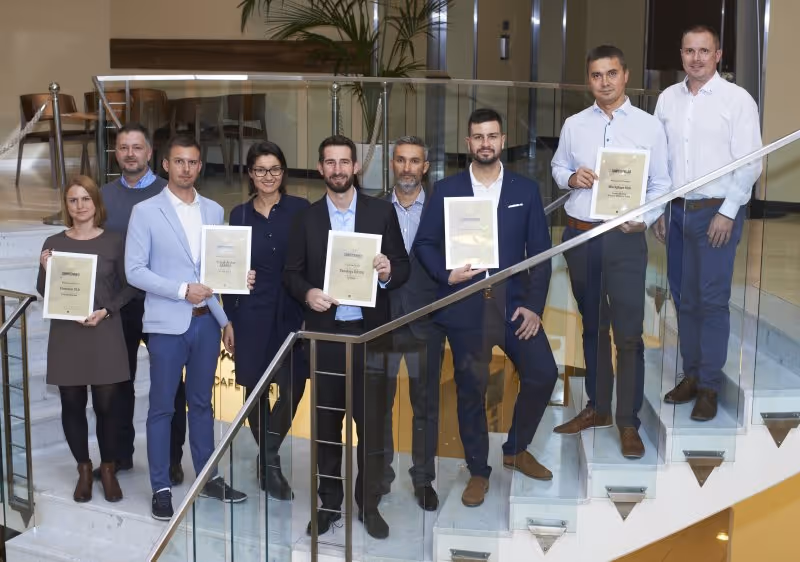 Skupina devíti lidí stojících na schodech, držících certifikáty v interiéru budovy.