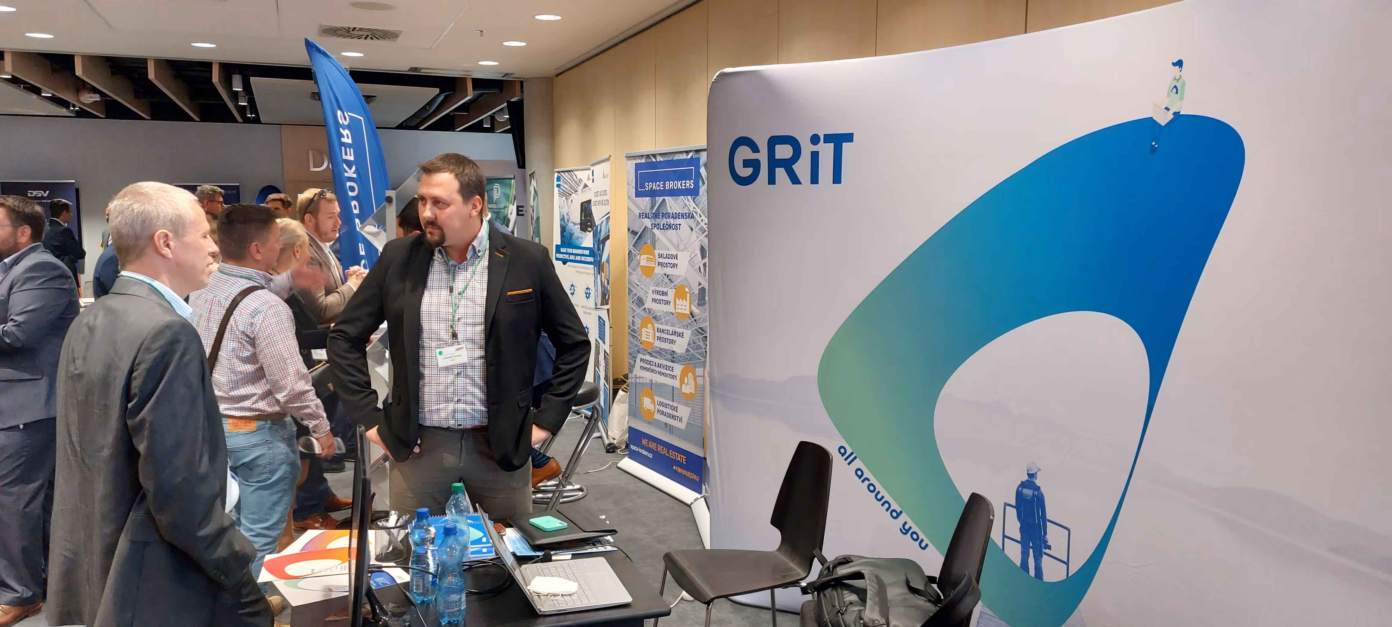 Lidé diskutují u stánku GRIT na konferenci s propagačními materiály a obrazovkou.