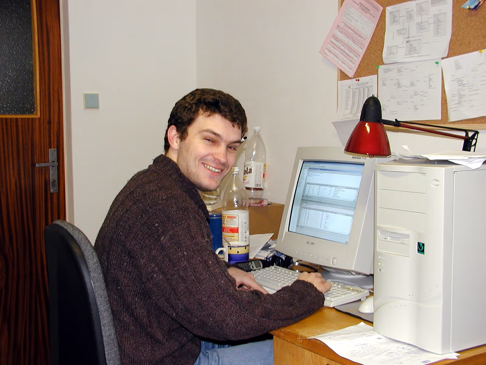 Muž se směje sedící u starého počítače s CRT monitorem v kanceláři s papíry na tabuli.