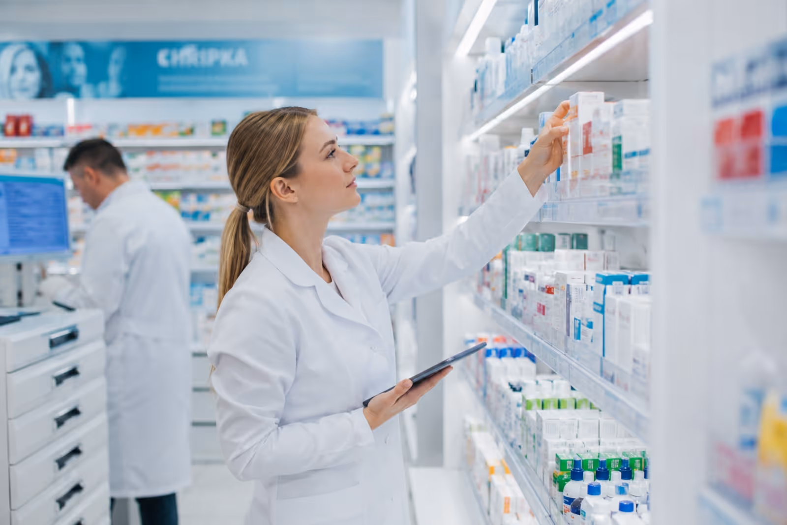 Farmaceutka v bílém plášti vybírá lék z police v lékárně držíc tablet.