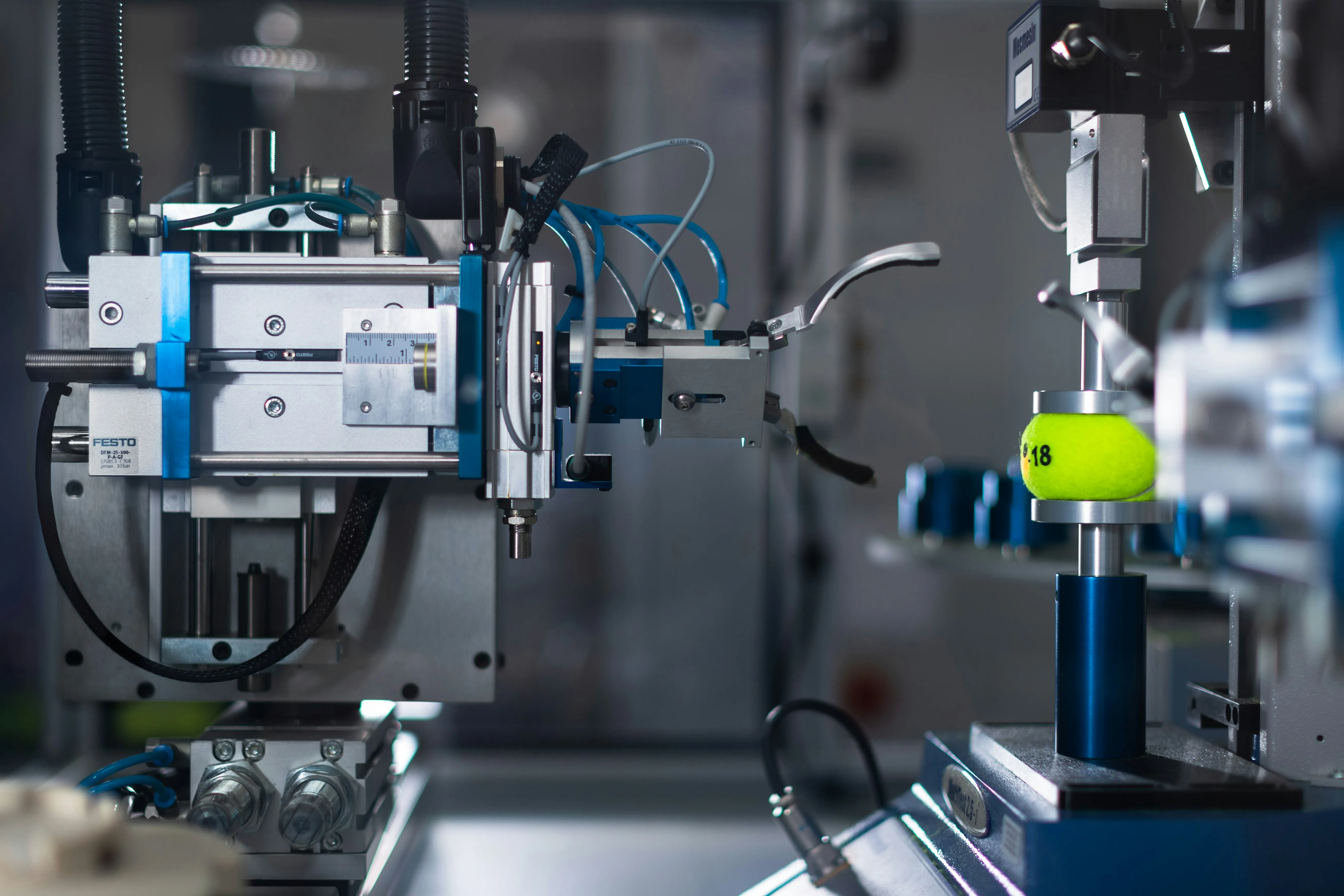 Industrial machine holding a yellow tennis ball for automated testing or processing in a lab environment.