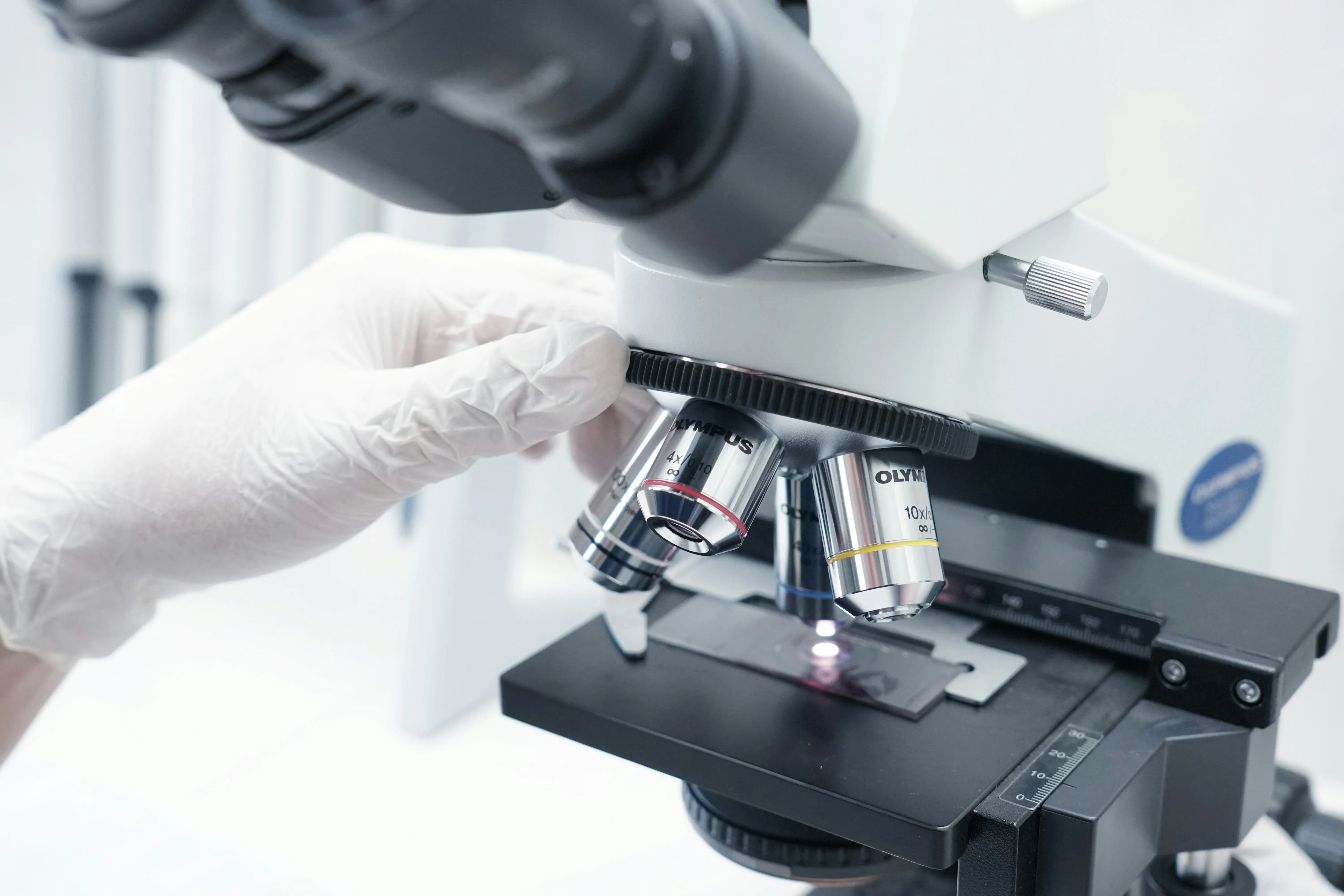 Gloved hand adjusting an Olympus microscope viewing a sample slide in a lab setting.