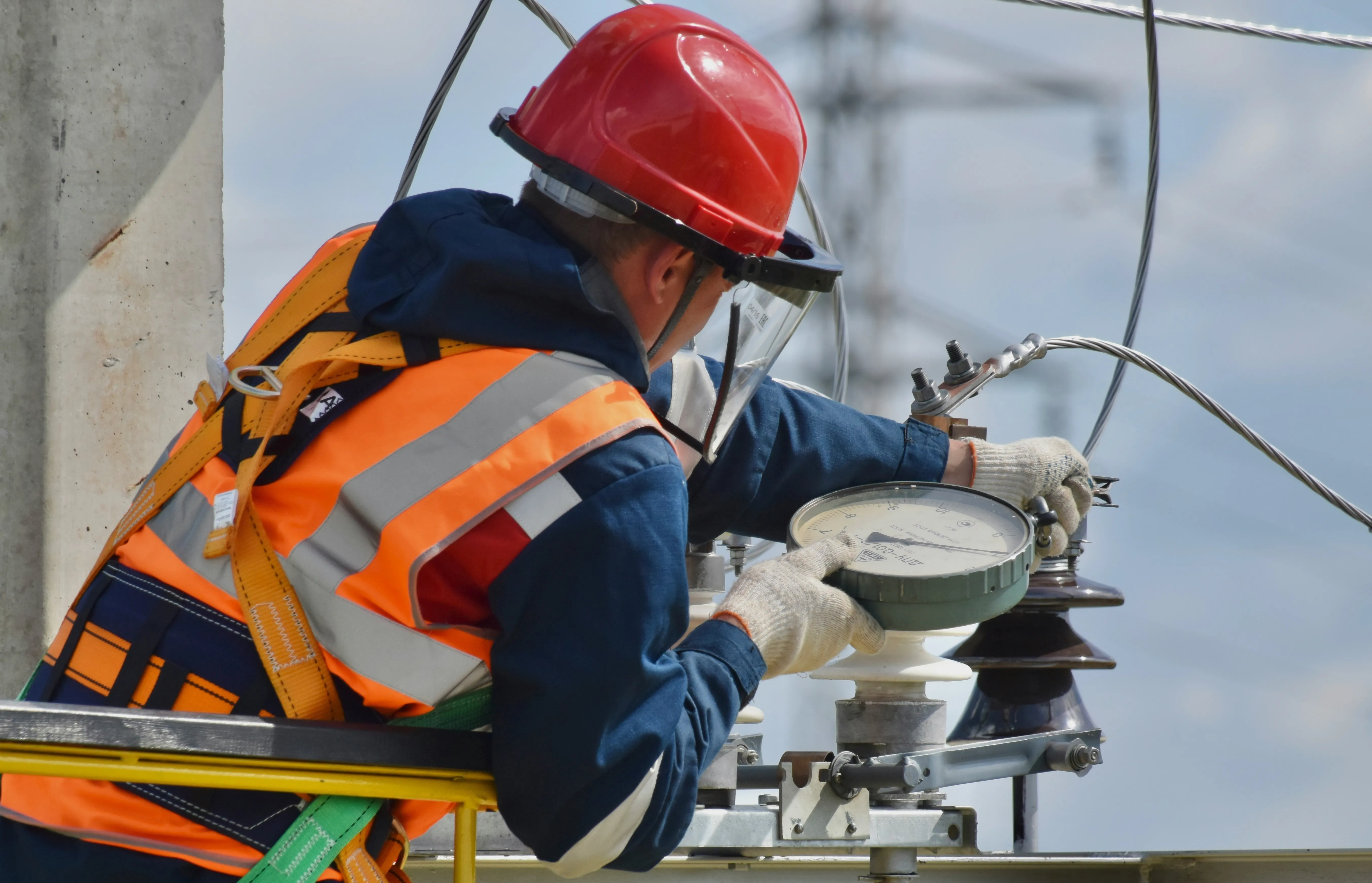 Utility worker in safety gear checking a gauge on electrical power equipment outdoors.