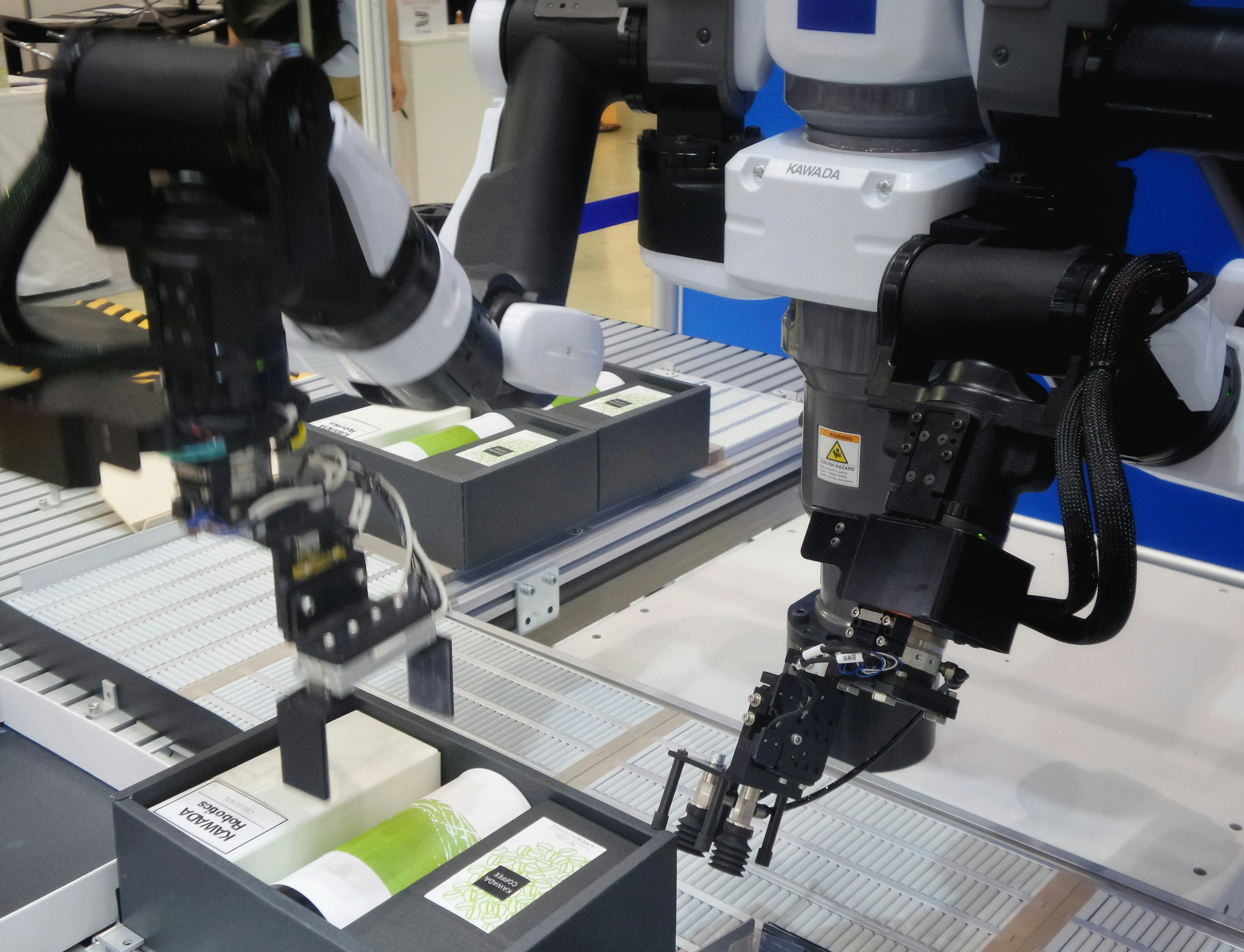 Robotic arms organizing packaged products labeled Kawada Robotics on a conveyor belt in a factory.