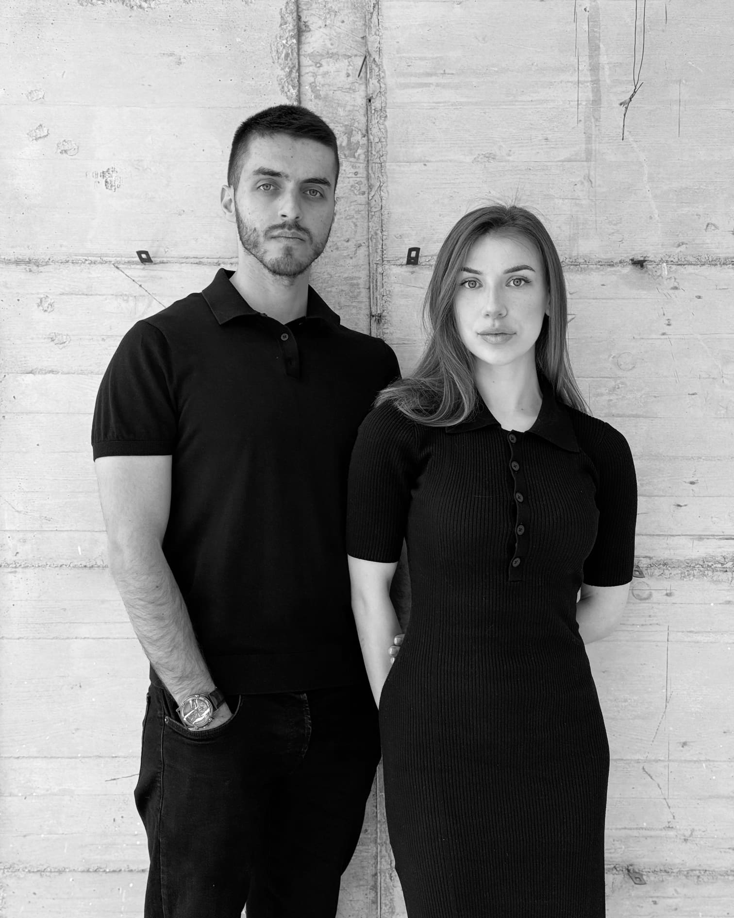 Black and white photo of a man and woman standing against a concrete wall, both wearing black outfits.