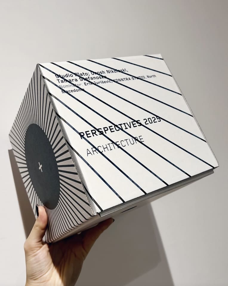 Hand holding a white cube with black striped design and text about Perspectives 2025 Architecture.