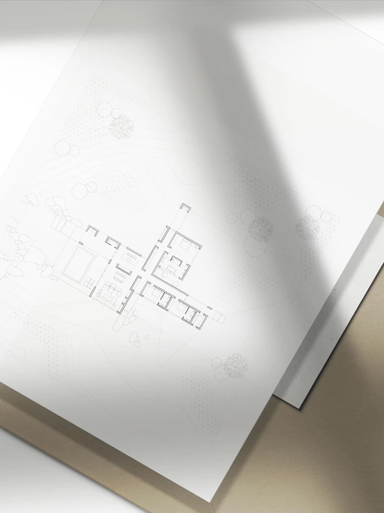 Architectural floor plan drawing on white paper with a shadow cast, showing a building layout with rooms and surrounding landscape elements like trees and rocks depicted by dotted lines and circles.