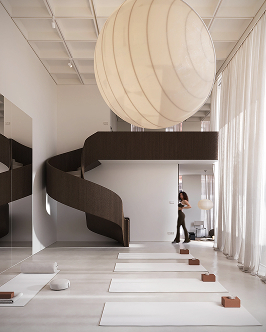 Bright, minimalistic yoga studio with white mats aligned on the floor, a large spherical ceiling light, a dark spiral staircase on the left, and sheer curtains letting in natural light on the right, with a person standing near the wall in the background.