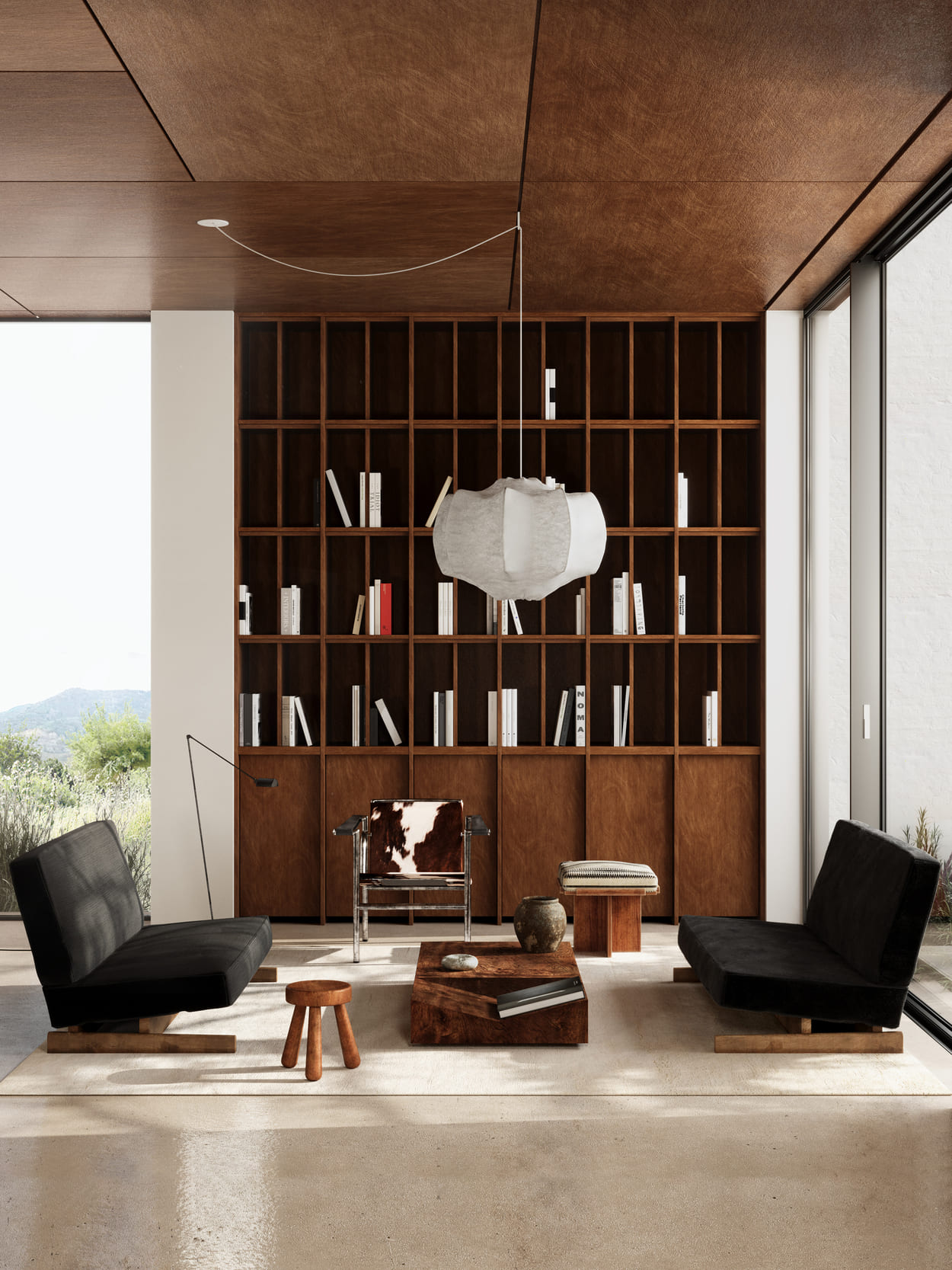 Modern living room with two black sofas facing each other on a beige rug, a wooden coffee table with books and pottery, a cowhide armchair, a small wooden stool, and a low wooden bench with a cushion, all set against a tall dark wooden bookshelf filled with spaced white and red books, under a wooden paneled ceiling with a white hanging pendant lamp, next to large floor-to-ceiling windows showing greenery outside.