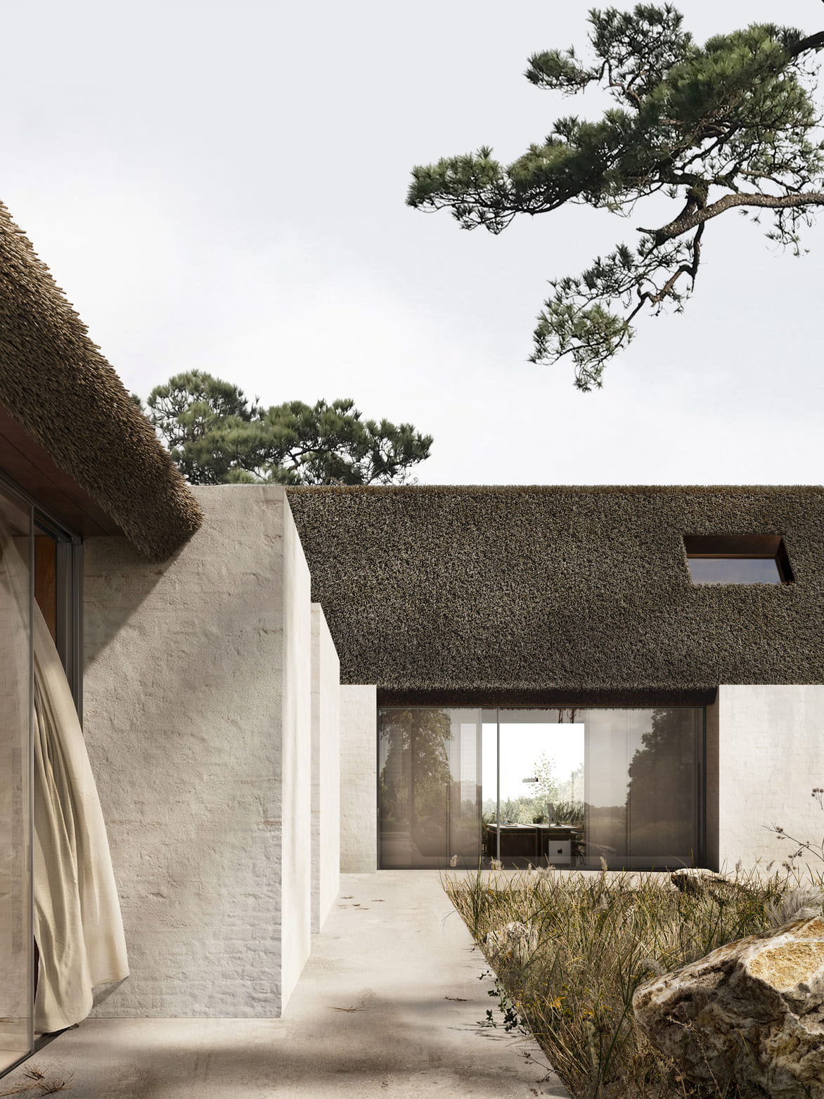 Modern house with white textured walls and a dark thatched roof, featuring large glass sliding doors opening to a courtyard with tall grass and rocks under an overcast sky with pine tree branches overhead.