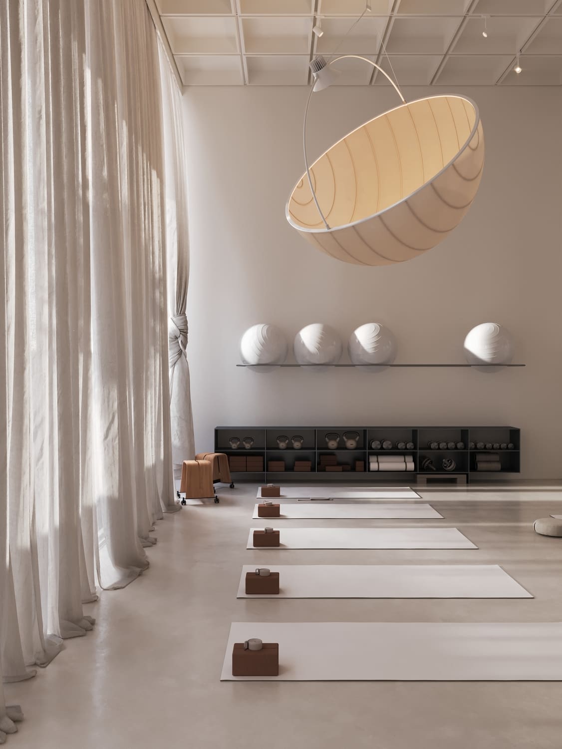 Minimalist yoga studio with five white mats lined up on a polished floor, each with a brown yoga block and strap at one end, large sheer curtains on the left, a wall shelf holding kettlebells and yoga props, and four large exercise balls on a floating shelf above.