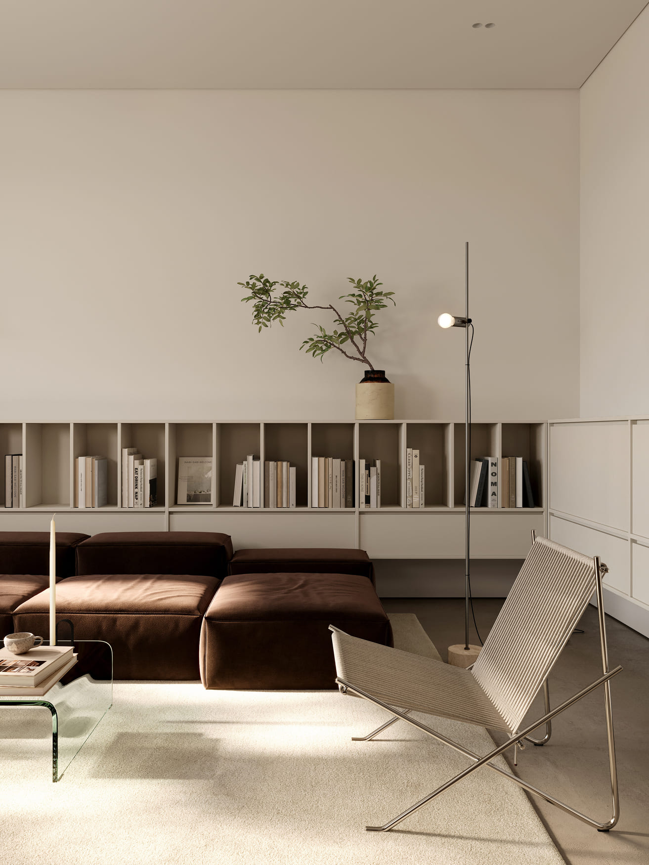 Minimalist living room with a brown modular sofa, a beige modern lounge chair with metal frame, a glass coffee table holding a candle and books, a long white wall-mounted bookshelf filled with books, a potted branch plant on the shelf, and a tall floor lamp with an exposed bulb, all set on a light beige carpet.