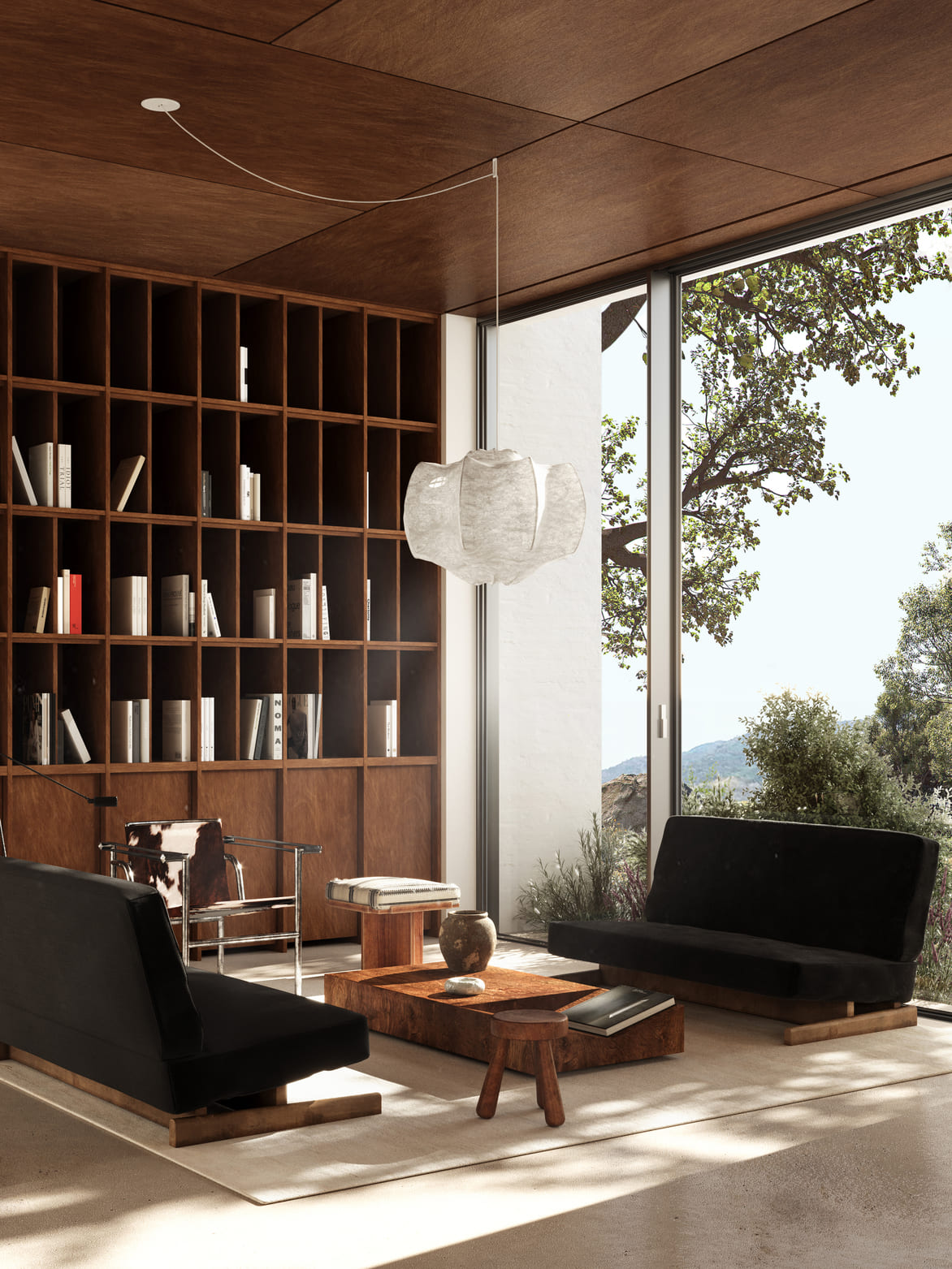 Modern living room with large floor-to-ceiling windows showing trees outside, two black sofas on a beige rug, a low wooden coffee table with decorative items, a small wooden stool, a cowhide armchair, and a tall wooden bookshelf filled with books against a wooden-paneled wall and ceiling.