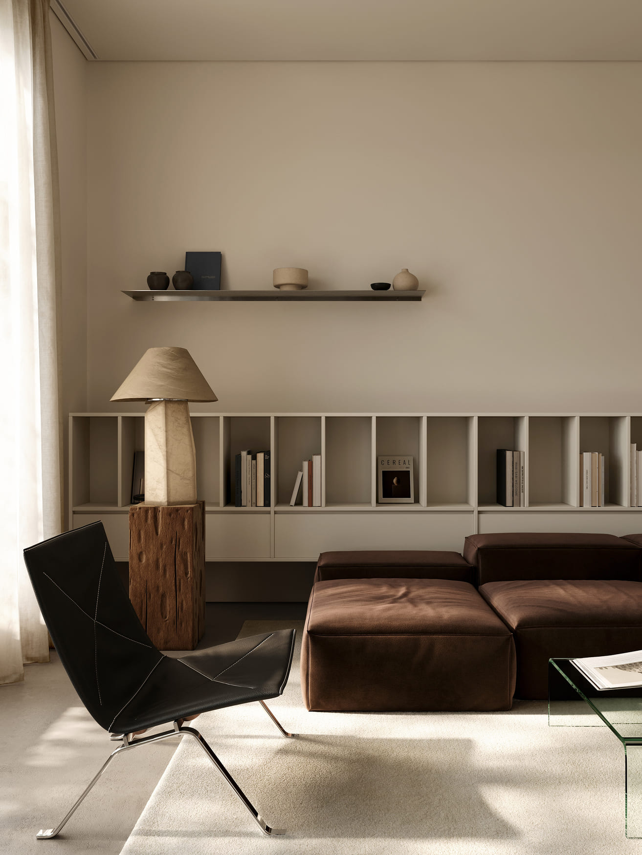 Minimalist living room with a black leather lounge chair with metal legs, a brown modular sofa, a wooden lamp with a beige shade on a wooden stump base, a white shelving unit with books, and a floating shelf holding small pottery and a book, all bathed in soft natural light from sheer curtains.