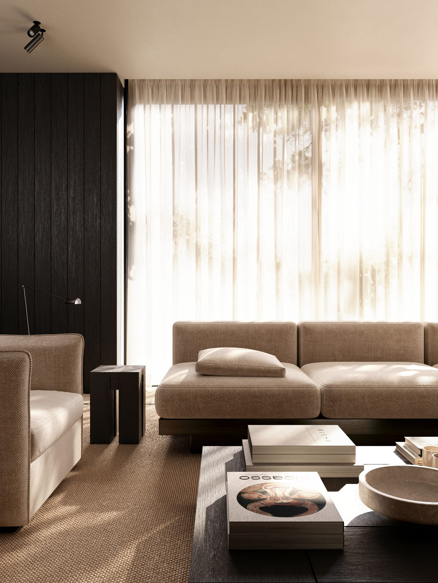 Sunlit modern living room with beige cushioned sofa and armchair, dark wooden coffee table with stacked books and a decorative bowl, textured brown carpet, sheer curtains covering large windows, and a dark wood accent wall with a floor lamp.