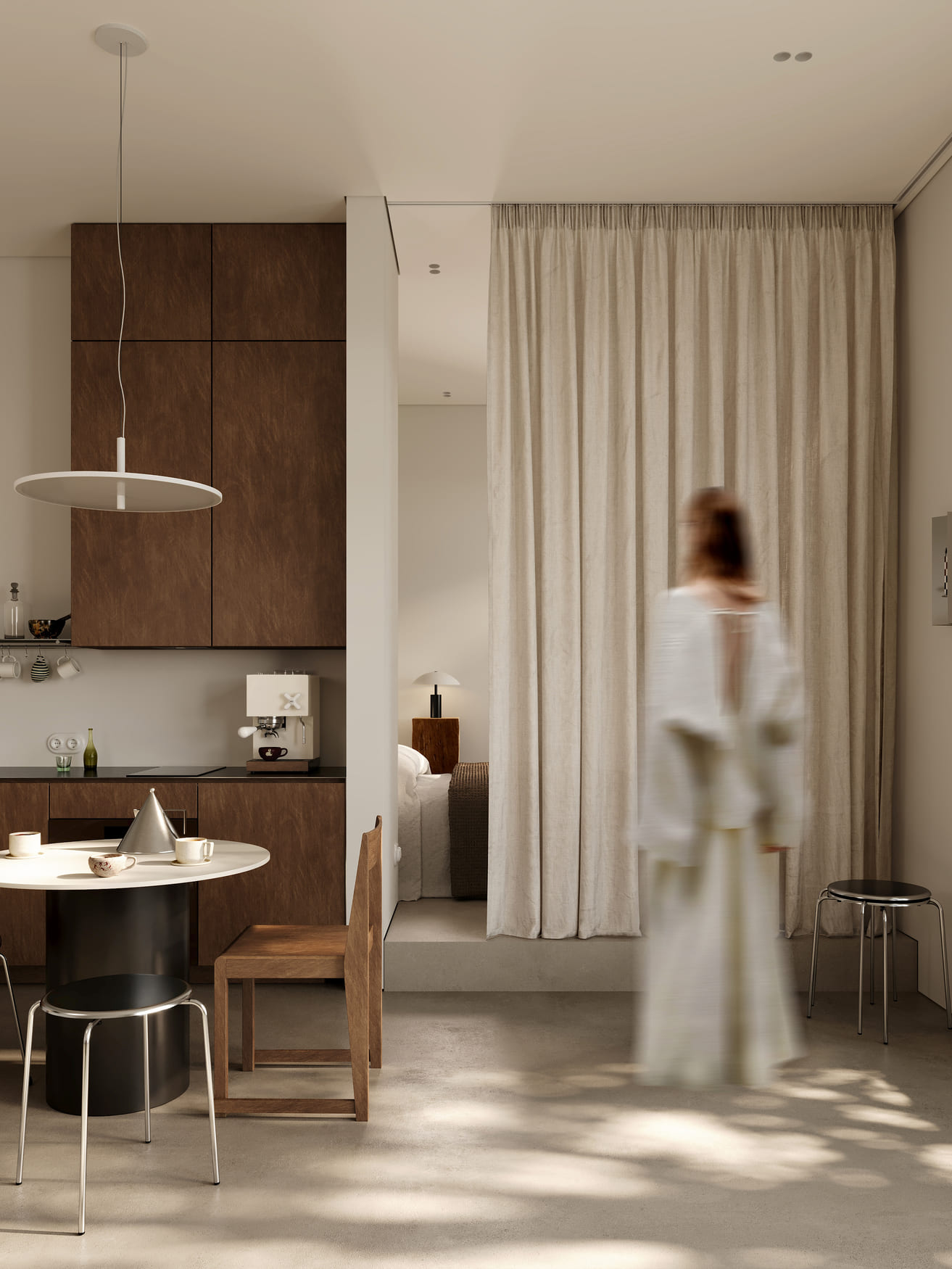 Minimalist beige and brown kitchen-dining area with modern cabinets, round table with cups and a cone-shaped object, wooden chair and metal stools; beige curtain partially conceals a bedroom with a lamp and bed; a blurred figure in a white dress stands nearby.