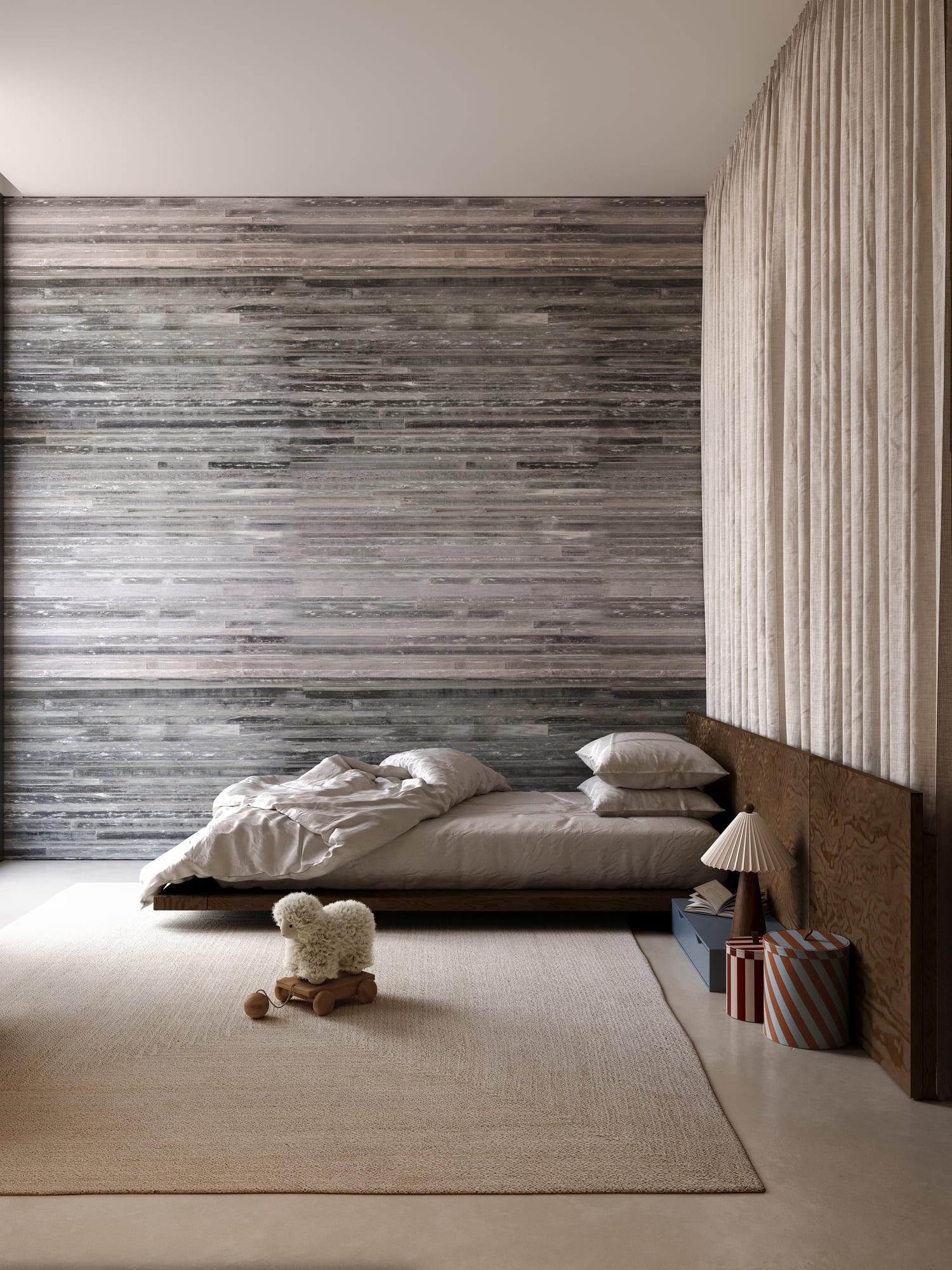 Minimalist bedroom with a low wooden bed featuring beige bedding and pillows, a textured grey wooden wall behind, floor-to-ceiling beige curtains on the right, a beige rug on the floor, and a plush sheep toy on wheels placed in front of the bed, accompanied by two striped storage boxes and a table lamp with a pleated lampshade beside the bed.