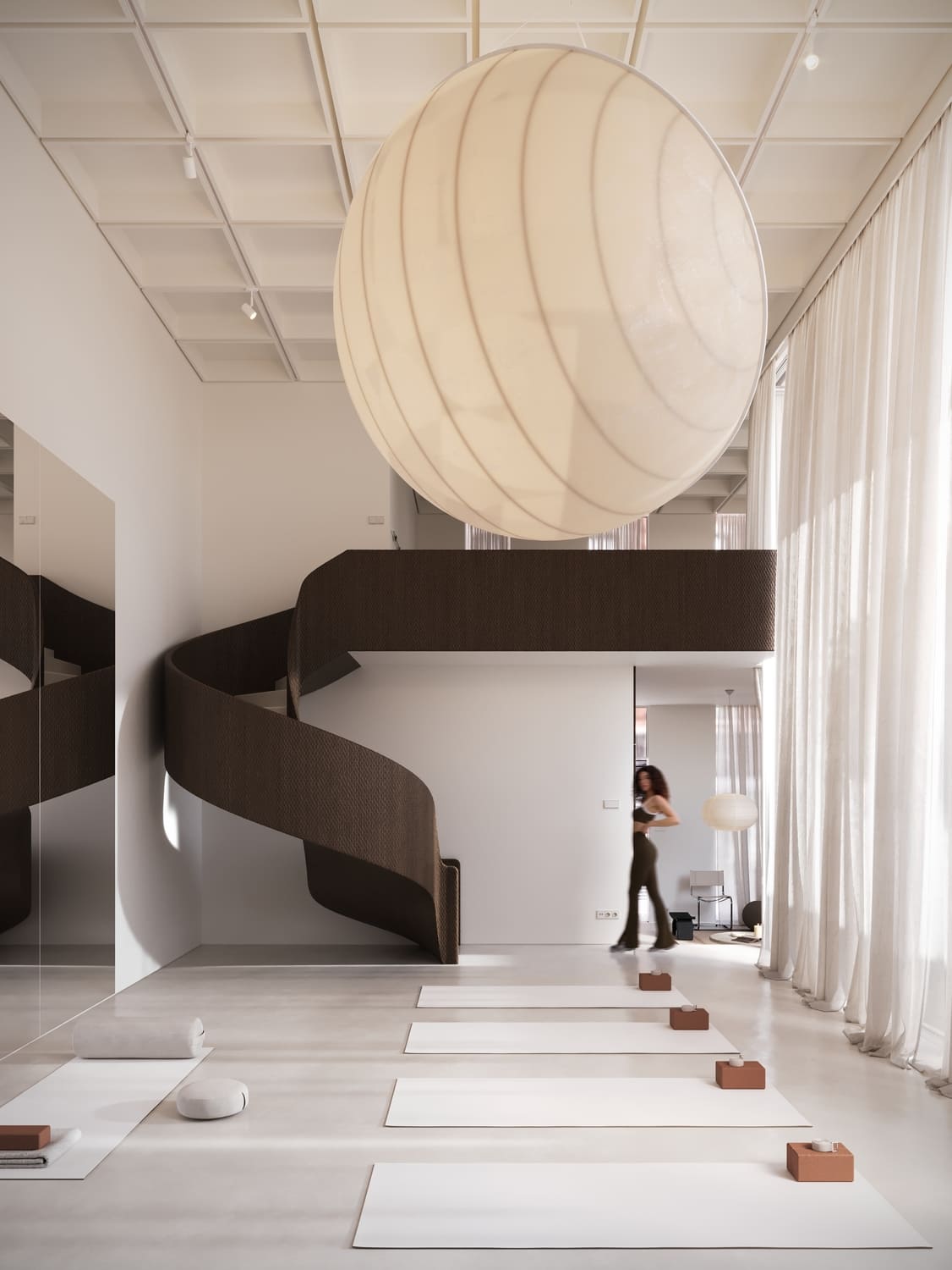 Minimalist interior with white floor mats aligned on light flooring, each paired with a small brown block and candle, featuring a large hanging round lamp, a dark brown spiral staircase, tall sheer white curtains, and a blurred figure walking near a white wall.