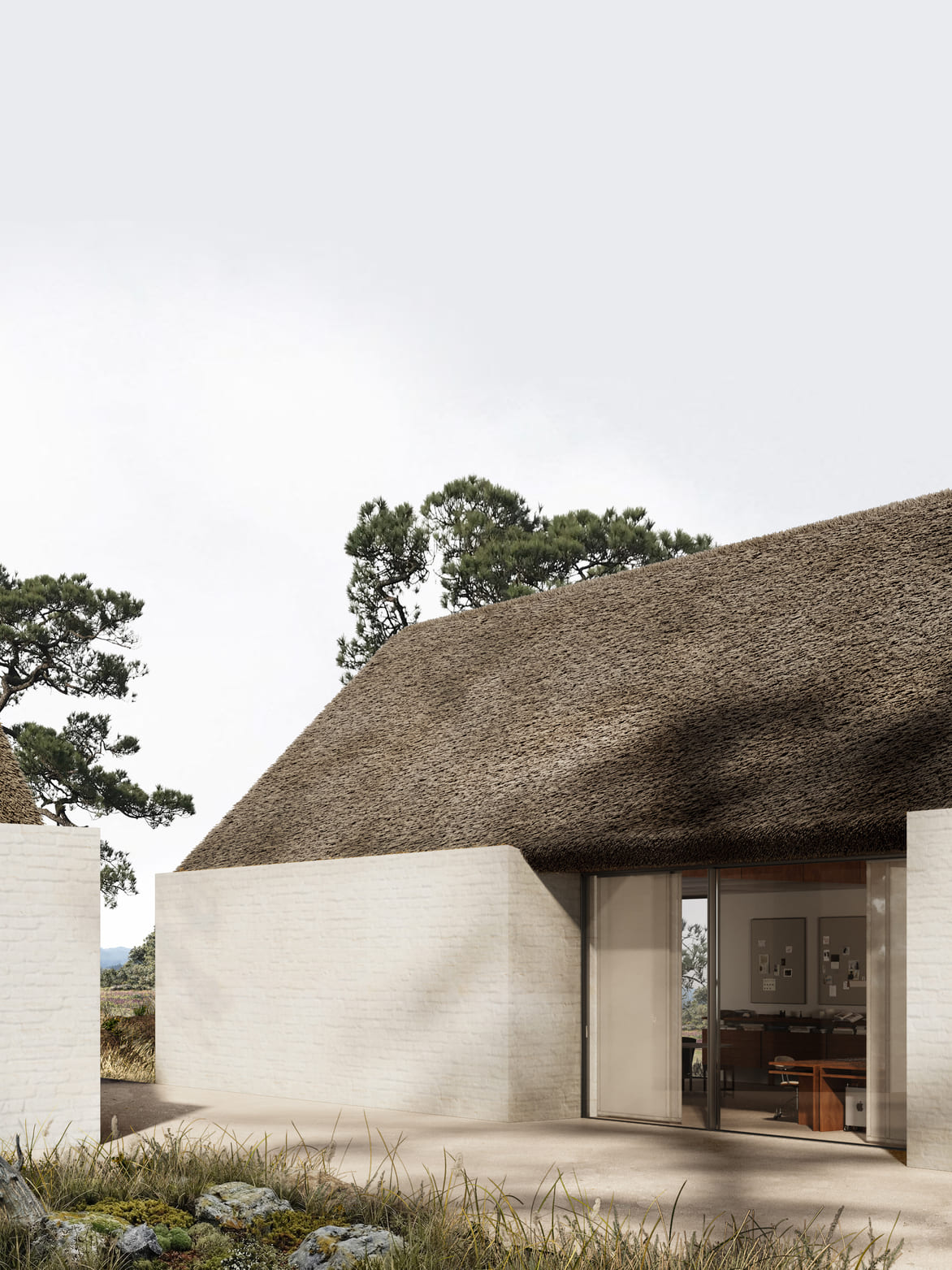 Modern building with white brick walls and a large thatched roof, featuring a glass sliding door revealing a wooden desk and office chair inside, surrounded by natural grass, rocks, and trees under a clear sky.