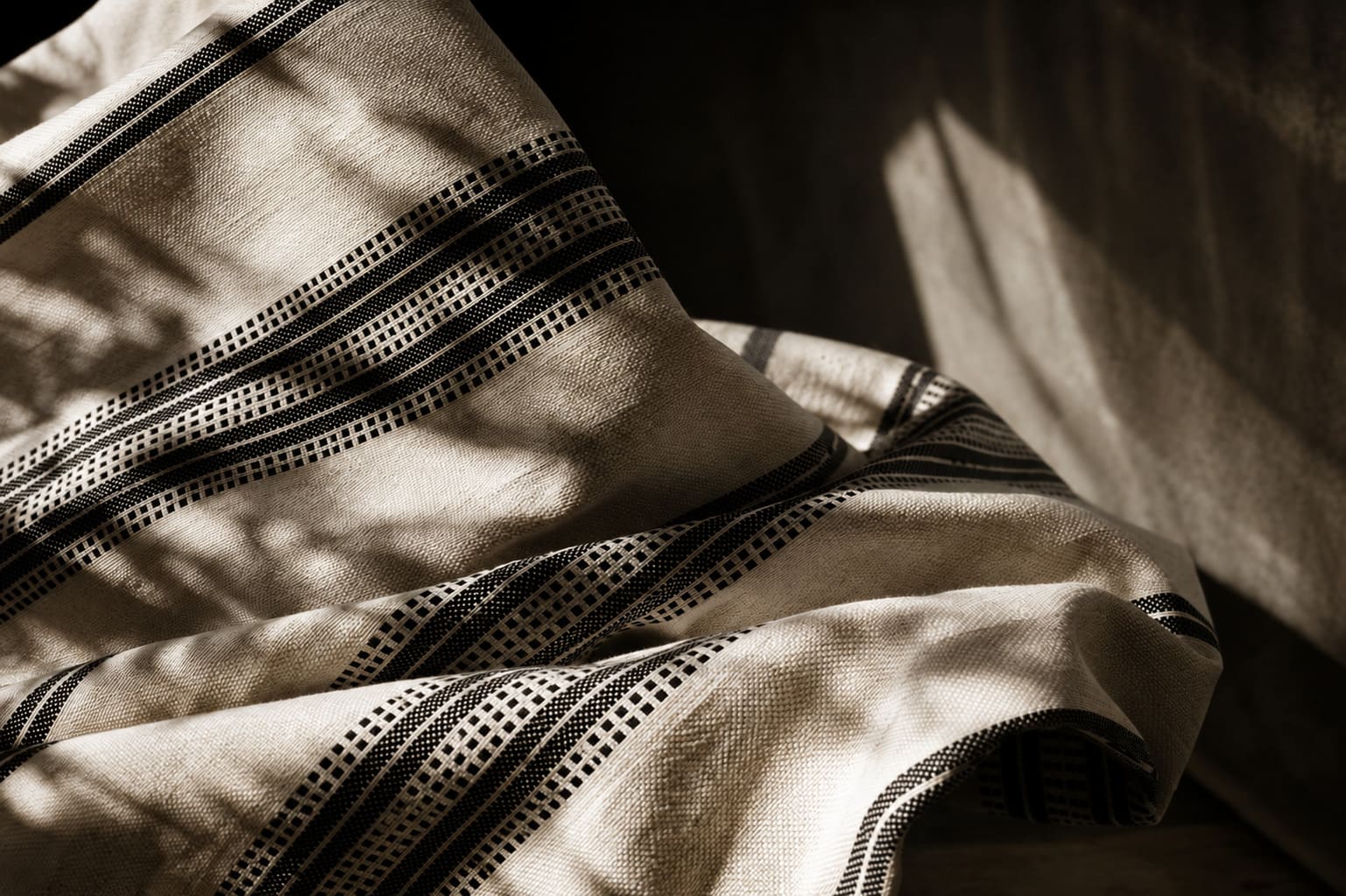 Close-up of beige fabric with black geometric stripes, softly folded with shadows creating a textured pattern.