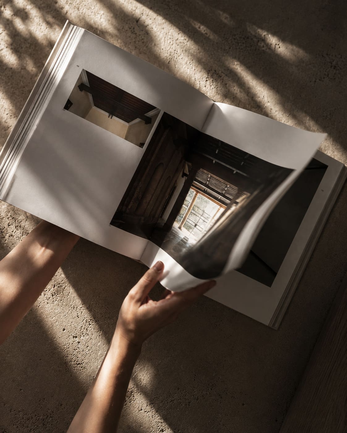 Person's hands flipping through a large open book with photographs of architectural interior spaces, on a textured surface with soft shadows.