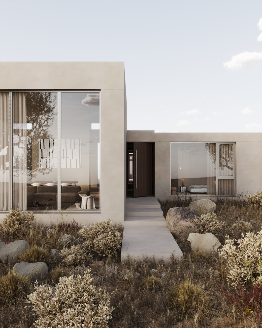 Modern one-story house with beige exterior and large glass windows showing interior furniture; surrounded by dry grass, bushes, and large rocks with a concrete pathway leading to a dark wooden front door.