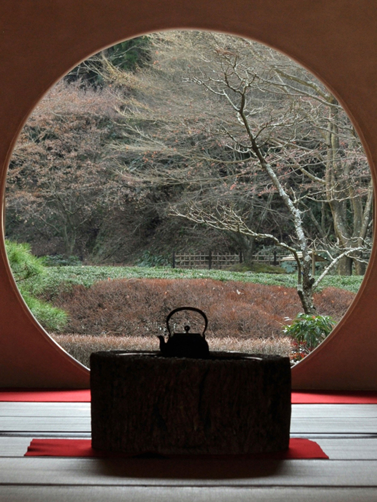 Silhouette of a traditional teapot placed on a low wooden block inside a room with a large circular window opening to a garden with bare trees and various bushes.